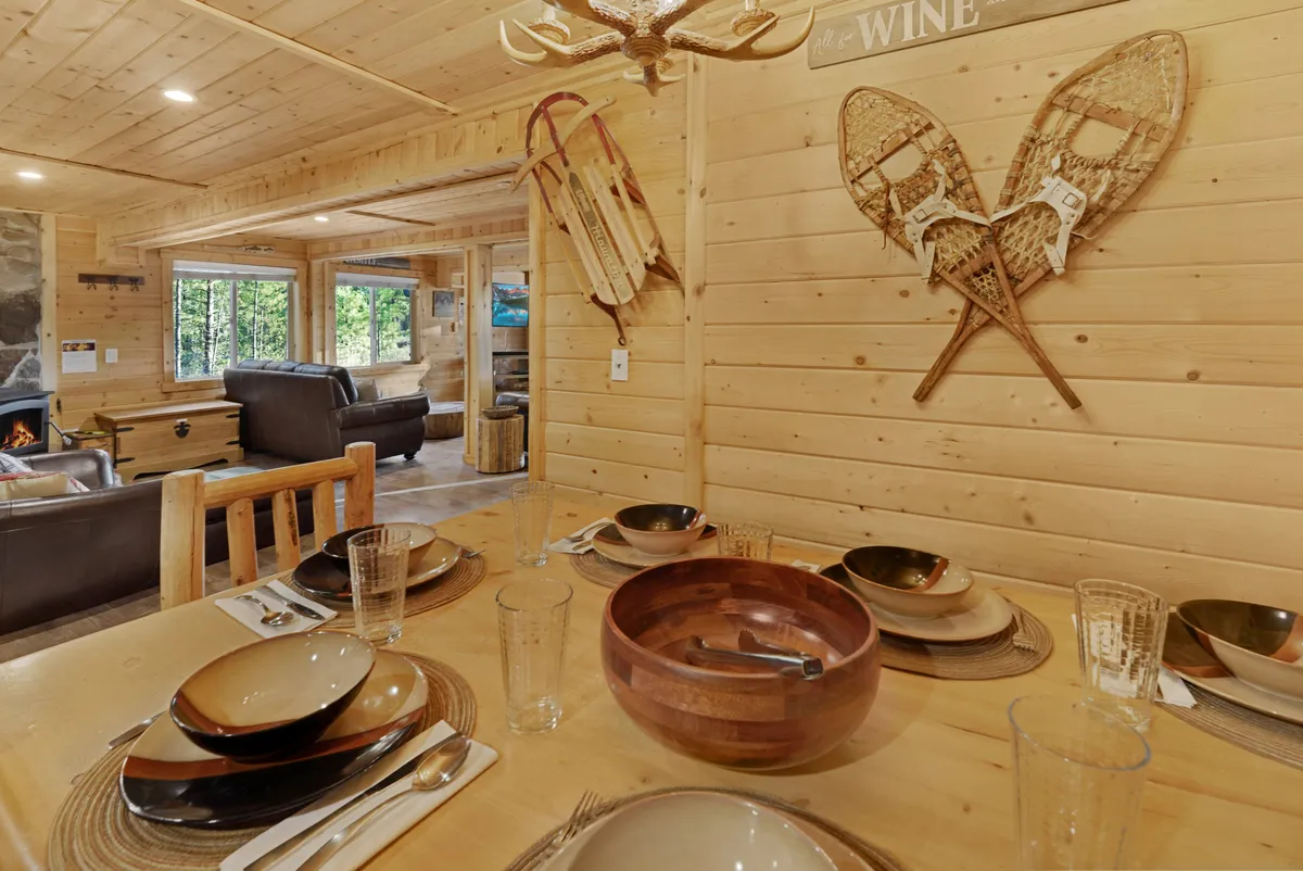 A rustic dining space with a large wooden table set with dishes and a decorative wooden bowl. Snowshoes hang on the wall as mountain-themed art, and a fireplace is visible in the background, tying together dining and lounging areas. Comfortable seating and warm lighting evoke a welcoming atmosphere for shared meals and conversation.
