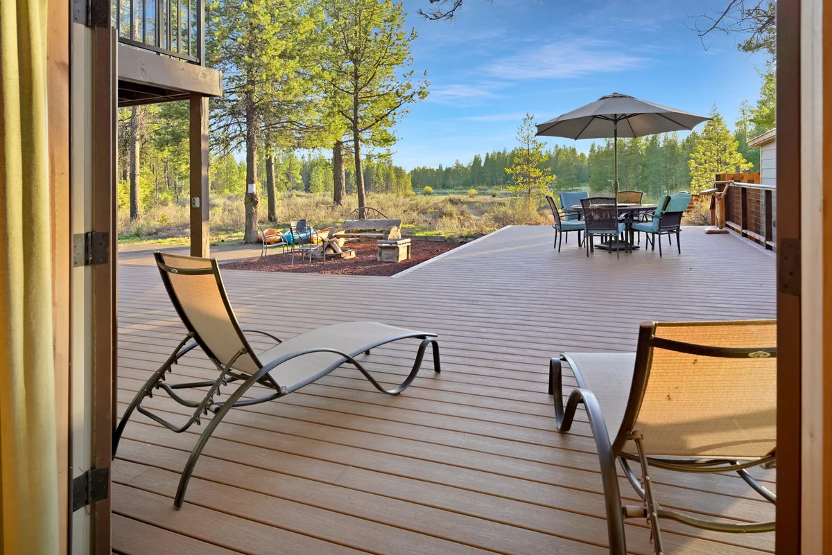 This image shows an inviting outdoor deck with lounge chairs, a dining table under a large umbrella, and a fire pit, all set against a backdrop of trees. The deck is elevated, providing a scenic vantage point over the surroundings, ideal for relaxing, dining, or gathering around the fire.