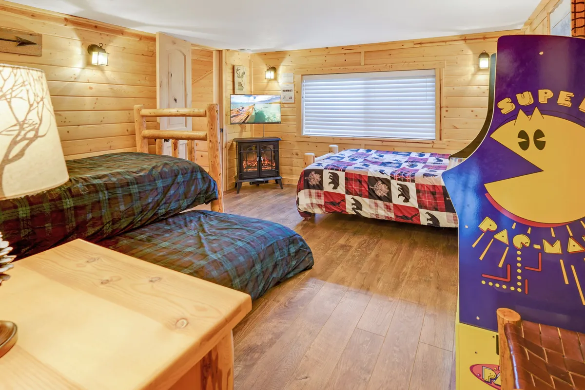 A playful, family-friendly bedroom featuring bunk beds, a queen bed, a Pac-Man arcade game, fireplace, and TV. The room is spacious and versatile, with options for sleeping, relaxing, and entertainment for all ages.