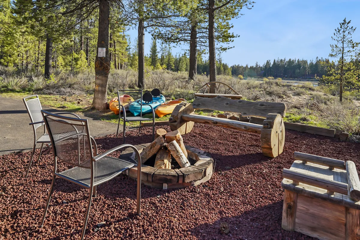 A welcoming fire pit area with wooden benches and chairs sits beside colorful kayaks, all overlooking a lake or river. The scene suggests both daytime fun on the water and communal evenings spent stargazing by the fire, with plenty of space for family and pets to play safely.