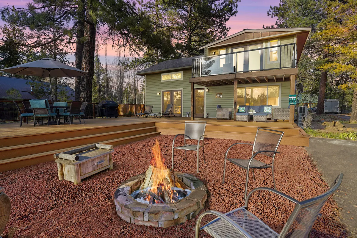The image shows the inviting exterior of a single-family cabin nestled among towering trees along the Big Deschutes River. The focus is on a spacious deck area, cozy outdoor seating, and a stone fire pit where guests can gather. The scene captures an atmosphere of relaxation, with people enjoying time together around the fire, dining outdoors, and taking in the natural beauty of the surroundings.