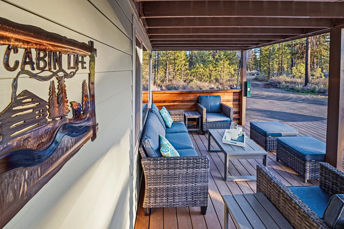 A cozy cabin deck area with a 'Cabin Life' sign, comfortable wicker seating, a coffee table, and wooden decking. The scene looks out onto nature, inviting guests to relax, socialize, and enjoy the outdoors right at the property.