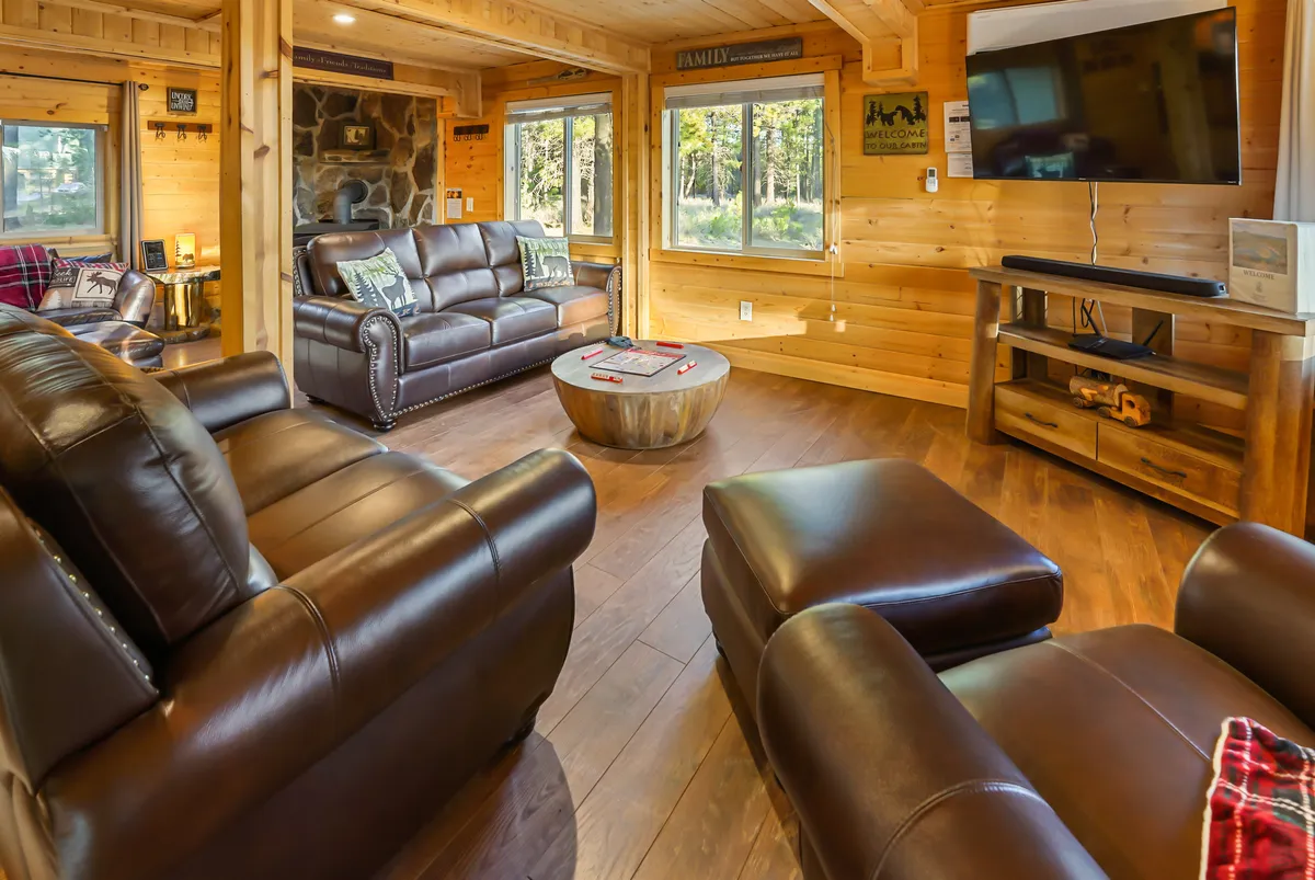 A spacious living area featuring leather sofas arranged around a coffee table, with a prominent stone fireplace and a large television. Expansive windows in the background frame woodland views, filling the room with natural light and connecting the indoors to the outdoors. The decor is warm and rustic, ideal for gatherings, movie nights, or cozy fireside relaxation.