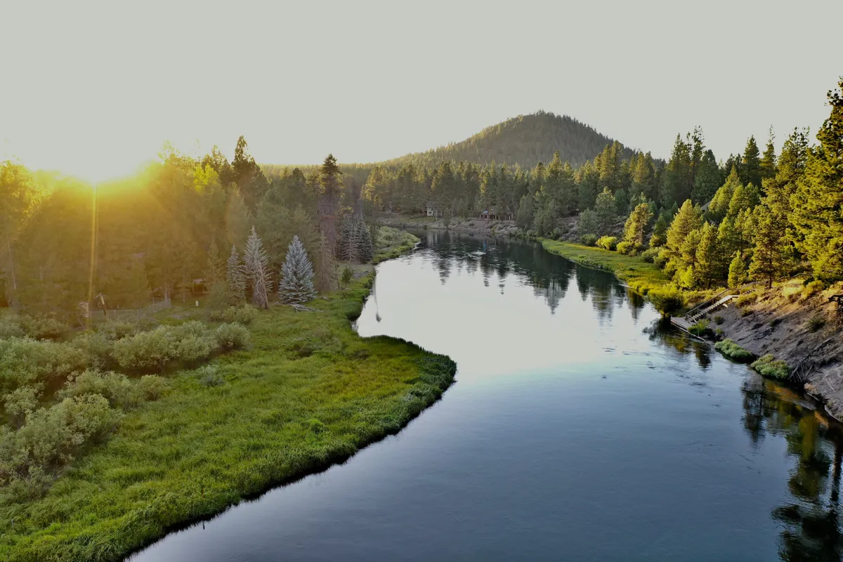 A stunning view of a river winding past mountains and through a forest of trees, with the warm glow of sunset reflecting on the water. This picturesque scene sets a serene, adventure-ready mood and evokes a sense of vast outdoor possibilities.