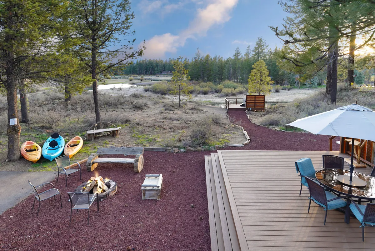 The exterior setting features kayaks resting near a fire pit, a relaxed outdoor dining area, and a pathway leading directly down to the water. The surrounding trees and open sky evoke a sense of relaxation and adventure, inviting families to gather for campfires, embark on kayaking trips, or simply enjoy meals with a view.