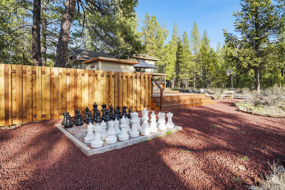 This playful exterior scene features a large outdoor chess set on a manicured lawn, a wooden deck area, a basketball hoop, and a fenced yard visible in the background. The space is ideal for children, pets, and family fun.