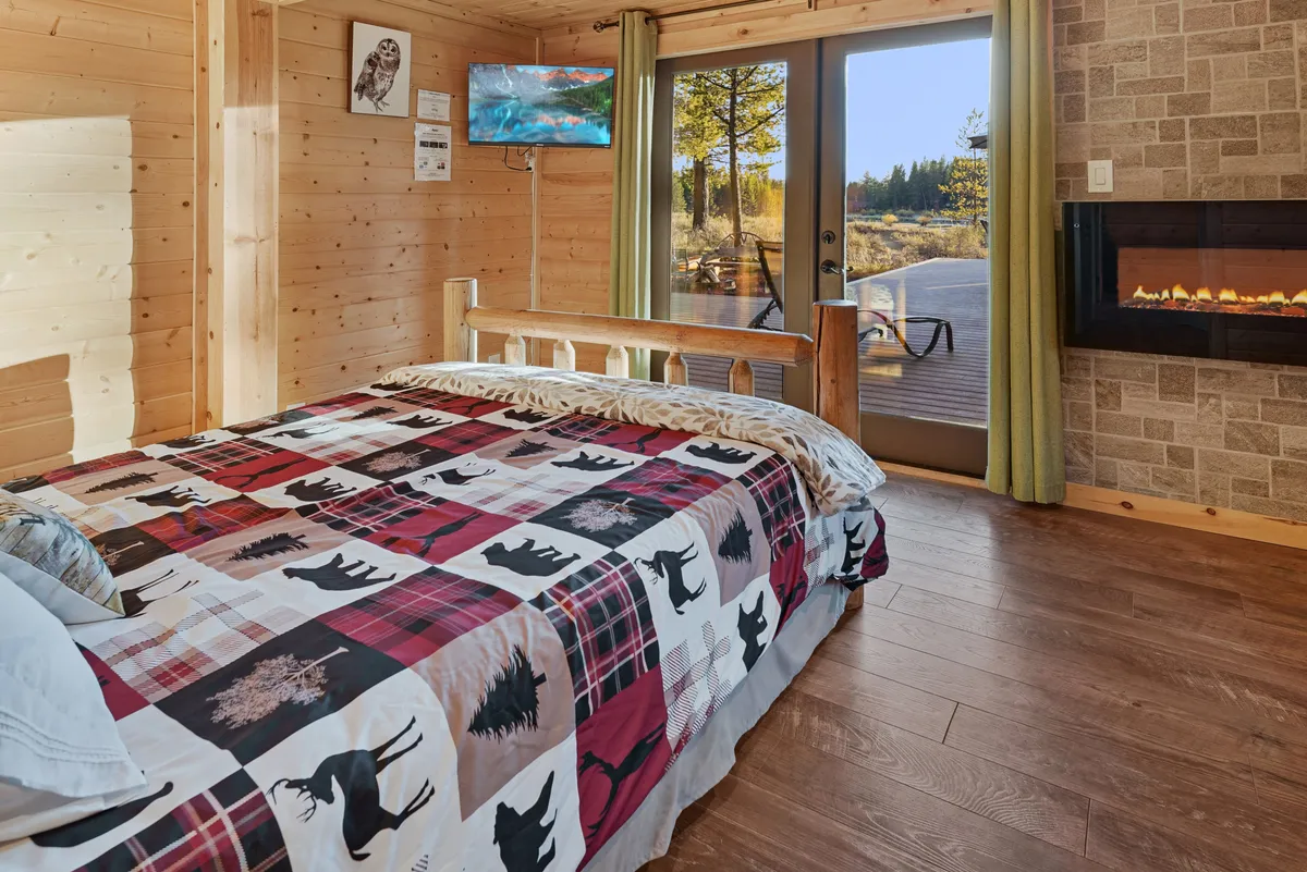 A king-sized bed adorned with a patterned quilt faces a cozy fireplace, with a large window offering views of the outdoors and a TV mounted on the wall. The space feels spacious, warm, and invites relaxation, suggesting access to a private deck just out of sight.